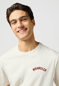 Hvid bomulds t-shirt med en buet "WRANGLER" grafik i bordeaux broderi på brystet. Klassisk rund halsudskæring og korte ærmer.