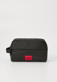 Svart nylon toalettmappe med glidelås på toppen, flat design, liten håndtak og en fremtredende rød rektangulær logo-patch med teksten "HUGO."