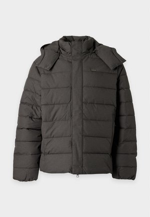 Homme debout, mains dans les poches, portant une veste matelassée vert foncé, un pantalon noir et des baskets blanches sur un fond clair uni.