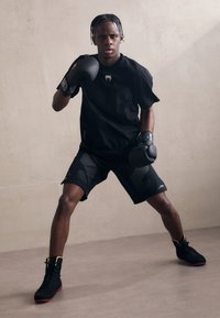Černé sportovní tričko s krátkým rukávem a logem, doplněné černými síťovanými šortkami s akcenty. Černé boxerské rukavice a boty s žlutými detaily.