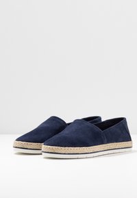 Scarpe slip-on in suede blu navy con suola in corda di juta, dotate di una base piatta in gomma bianca e un design aperto sul retro.