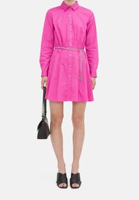 Abito button-down fucsia con maniche lunghe, vita arricciata, cintura in catena argentata, abbinato a una borsa nera e sandali con tacco neri aperti.