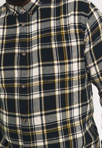 Rutiga skjortan med knappslå har en färgkombination av marinblått, vitt och gult, med en borstad textur och en standardkrage.