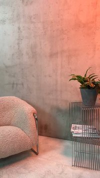 Gemütlicher beiger strukturierter Sessel neben einem Metall-Draht-Beistelltisch mit gestapelten Magazinen und einer Topfpflanze vor einer Betonwand.