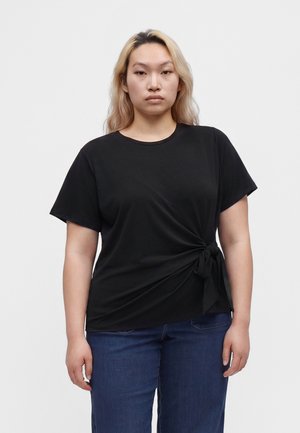 Jeune femme aux longs cheveux blonds porte un haut noir à manches courtes avec un nœud sur le côté et un jean bleu, debout devant un fond clair uni.