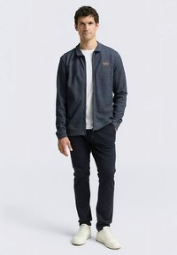 Herenjack met ritssluiting in donkergrijze stof, voorzien van een opstaande kraag, voorzakken en een geborduurd logo. Draagt een wit overhemd en marineblauwe broek.