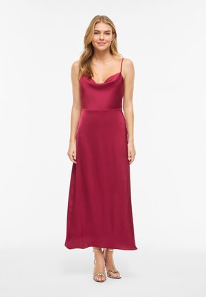 Naine seisab valgel taustal, kandes varrukateta, põrandani ulatuvat punast satiinist kleiti, millel on õhukesed sangad, ning kuldseid avatud varvastega kontsakingi.