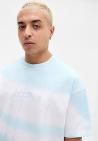 Jong persoon met kort gebleekt haar, die een blauw-wit tie-dye T-shirt draagt met een subtiel geborduurd logo op de borst.
