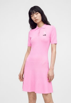 Femme portant une robe polo rose à manches courtes avec une jupe côtelée et un petit logo lapin blanc indiquant "Love Moschino" sur la poitrine.