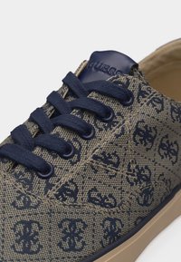 Κοντινό πλάνο ενός αθλητικού παπουτσιού με ναυτικό μπλε και μπεζ μοτίβο, με ναυτικά μπλε κορδόνια και μπεζ σόλα, που διαθέτει ανάγλυφο λογότυπο στη γλώσσα.