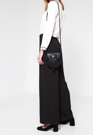 Nainen pukeutuneena valkoiseen puseroon, mustiin leveälahkeisiin housuihin, mustiin korkokenkään ja mustaan olkalaukkuun, jossa on etutasku ja hapsu.