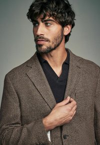 Brauner Hahnentrittmuster-Blazer mit strukturiertem Finish, kombiniert mit einem dunklen V-Ausschnitt-Pullover. Auffällige Details sind die einzelne Brusttasche und die Knöpfe.
