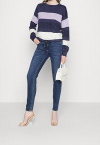 Donna che indossa jeans skinny blu scuro, un maglione navy con righe viola e bianche, sandali con tacco alto trasparenti e tiene in mano una piccola borsa bianca.