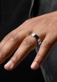 Metallring med en polert sølvfinish, flat design, glatt tekstur, båret på en hånd med naturlige negler, mot en mørk bakgrunn.