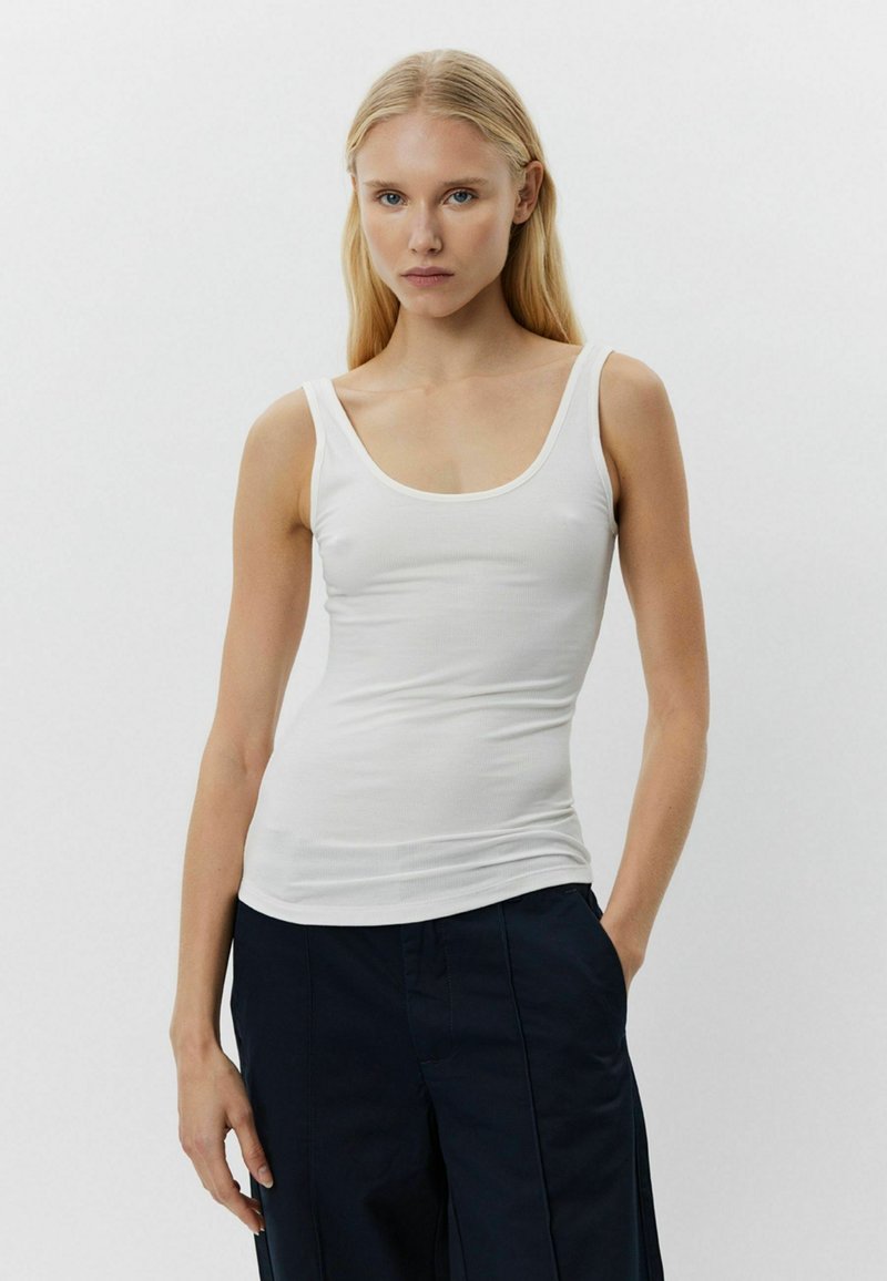 Fehér bordás trikó széles pántokkal és mélyen dekoltált nyakkivágással, testhezálló fazonú, sötét tengerészkék, bő szabású nadrággal párosítva.