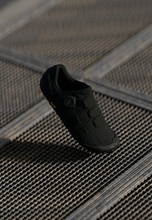 Černé lehké tenisky s texturovaným svrškem, elastickými tkaničkami a kruhovým zapínáním. Mají robustní podrážku s žlutým akcentem.