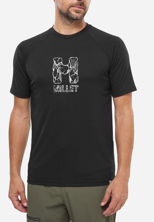 Mann trägt schwarzes Kurzarmhemd mit weißem abstraktem "M" und dem Schriftzug "MILLET", kombiniert mit olivgrünen Hosen vor einem einfarbigen Hintergrund.