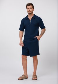 Marinefarbener Kurzarm-Polo mit Reißverschlusskragen, kombiniert mit passenden marineblauen Shorts. Der Stoff hat eine strukturierte Textur; Sandalen runden das Outfit ab.