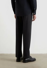 Schwarzes Hemd mit Knöpfen an den Manschetten, kombiniert mit dunklen, locker sitzenden Hosen und schwarzen Lederschuhen. Minimalistisches Design und glattes Material.