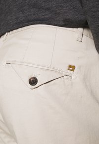Beige katoenen broek met een knoop achterzak en unieke driehoekige flapdetails. Heeft een klein gouden naaimachine-embleem.