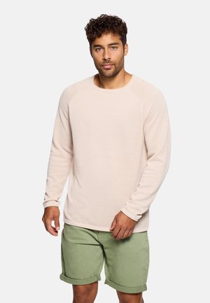 Hombre con cabello rizado que lleva un suéter beige de manga larga con textura y pantalones cortos verde oliva remangados, sobre un fondo liso.