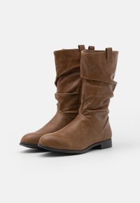 Bottes montantes marron en cuir synthétique avec un design décontracté, semelle plate noire, et détails à languette en haut. Texture lisse dans l'ensemble.