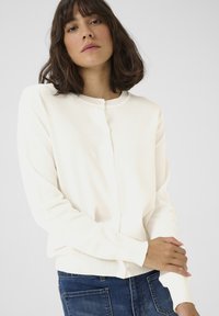 Biely pletený cardigan s okrúhlym výstrihom, diskrétnymi detailmi na gombíkoch a dlhými rukávmi, vystavený s modrými džínsami na neutrálnom pozadí.