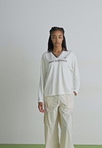 Weißes Langarmoberteil mit V-Ausschnitt und braunem "New Balance"-Logo, kombiniert mit beigen Cargo-Hosen, die seitliche Taschen und eine lockere Passform aufweisen.