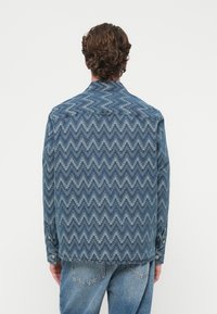 Jachetă din denim cu un model zigzag în nuanțe variate de albastru. Are guler, manșete cu nasturi și croială dreaptă la tiv.