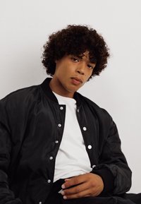 Μαύρο μπουφάν bomber με λευκά κουμπιά, φορεμένο πάνω από λευκό T-shirt. Σγουρά μαλλιά πλαισιώνουν μια ουδέτερη έκφραση. Απλό φόντο.