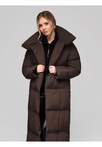 Lungo piumino marrone con un ampio colletto, caratterizzato da un motivo trapuntato e chiusure frontali con bottoni a pressione. Indossato sopra un maglione nero con zip.