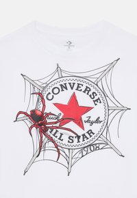 T-shirt en coton blanc avec un graphisme d'étoile rouge accompagné du texte "Converse Chuck Taylor", une illustration d'araignée et des accents de design en forme de toile d'araignée.