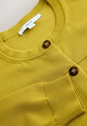 Dzeltens adīts kardigans ar apaļu kakla izgriezumu un divām brūnām pogām. Teksturēts audums ar ribotām detaļām aprocēs un kabatā.