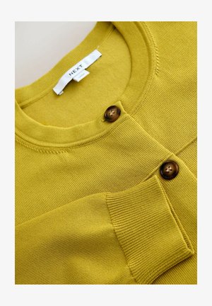 Żółty sweter zapinany na guziki z okrągłym dekoltem i dwoma brązowymi guzikami. Tkanina o fakturze z prążkowanymi wykończeniami na mankietach i kieszeni.