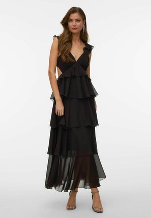 Naine kannab varrukateta musta kihilist maksikleiti volangide ja läbipaistvate paneelidega, millele on lisatud hõbedased kõrge kontsaga sandaalid ja kõrvarõngad.