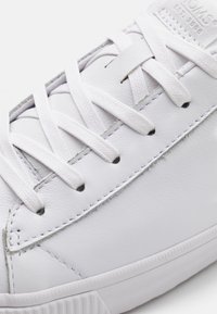 Baskets en cuir blanc avec des lacets tissés blancs, une texture lisse, des détails de coutures subtiles et une semelle en caoutchouc texturée.