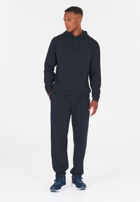 Sudadera de color azul marino con cordón, bolsillo frontal y pantalones de chándal a juego. El material es suave; presenta un ajuste holgado y una cintura elástica.