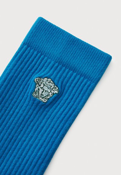 Blå ribbede sokker med en tekstureret overflade, der har et broderet emblem af et ansigt øverst. Blødt materiale, tætsiddende design.