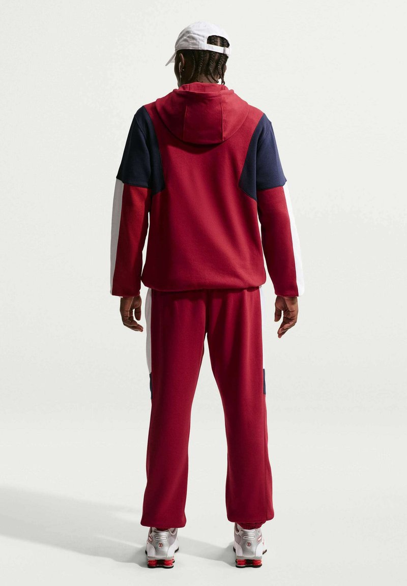 Persona che indossa una felpa con cappuccio rossa, blu navy e bianca con pantaloni abbinati, cappellino bianco e scarpe da ginnastica bianche, in piedi con le spalle rivolte verso uno sfondo uniforme.