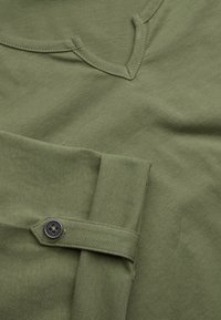 Olivegrünes Shirt mit V-Ausschnitt-Design, das über einen geknöpften Riemen am Ärmel verfügt. Der Stoff hat eine weiche, strukturierte Oberfläche.