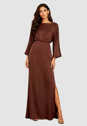 Donna che indossa un lungo abito di raso marrone con maniche lunghe, vita stretta e un alto spacco laterale, in piedi davanti a uno sfondo semplice.