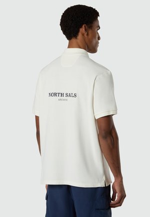 Hombre que lleva un polo blanco de manga corta con el texto "NORTH SAILS ARCHIVE" en la espalda, combinado con pantalones cortos cargo azul marino.
