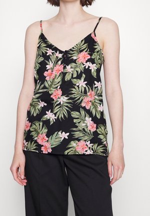 Kobieta w czarnej bluzce bez rękawów z różowo-zielonym tropikalnym kwiatowym nadrukiem i czarnych spodniach, stojąca na białym tle.