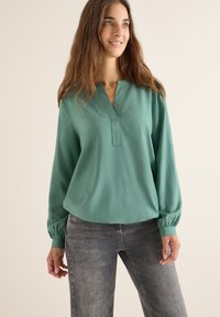 Türkise Bluse mit V-Ausschnitt, langen Puffärmeln mit elastischen Bündchen und lockerer Passform, kombiniert mit grauen Jeans. Glatte Stoffstruktur.