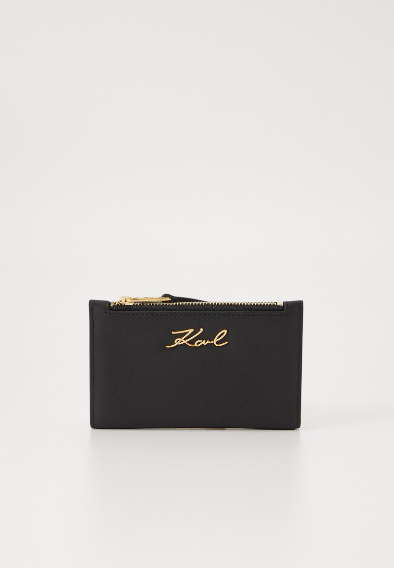 Crni novčanik izrađen od glatke kože, s zlatnim zatvaračem i utisnutim potpisom. Pravokutni oblik s minimalističkim dizajnom.