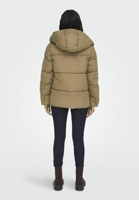 Hnědá péřová bunda s kapucí, s prošívaným designem a žebrovanými manžetami, v kombinaci s tmavými džínami a hnědými kotníkovými botami.