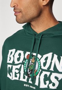 Felpa verde con la scritta "BOSTON CELTICS" in grande lettering bianco e un logo ricamato di un leprechaun che tiene un pallone da basket.