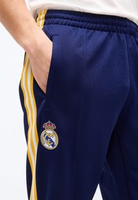Άτομο που φοράει αθλητικό παντελόνι ναυτικού μπλε χρώματος με κίτρινες και λευκές ρίγες στο πλάι και το λογότυπο της Ρεάλ Μαδρίτης, με το χέρι χαλαρά στην τσέπη.