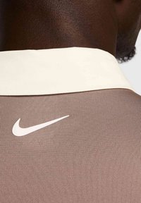 Brūna sporta krekls ar krēmkrāsas apkakli un baltu Nike logo aizmugurē. Audumam ir ribota tekstūra.