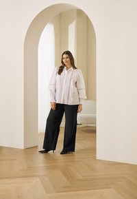 Weiße Bluse mit strukturierten Details, kombiniert mit schwarzen, weit geschnittenen Hosen und spitzen schwarzen Pumps. Lichtdurchfluteter Innenraum mit Holzboden.